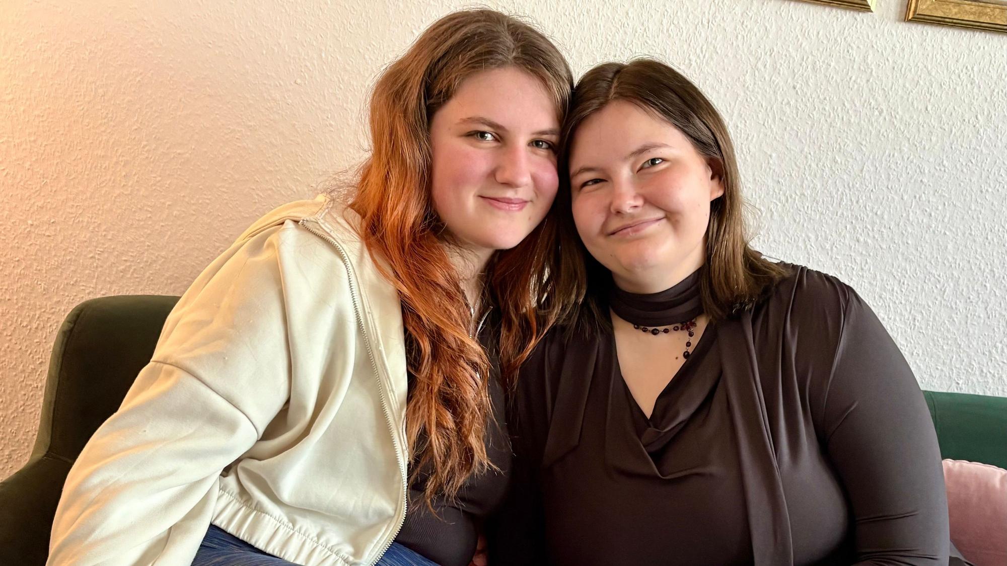 Die beiden jungen Frauen sitzen eng aneinander gekuschelt auf einem Sofa und lächeln.