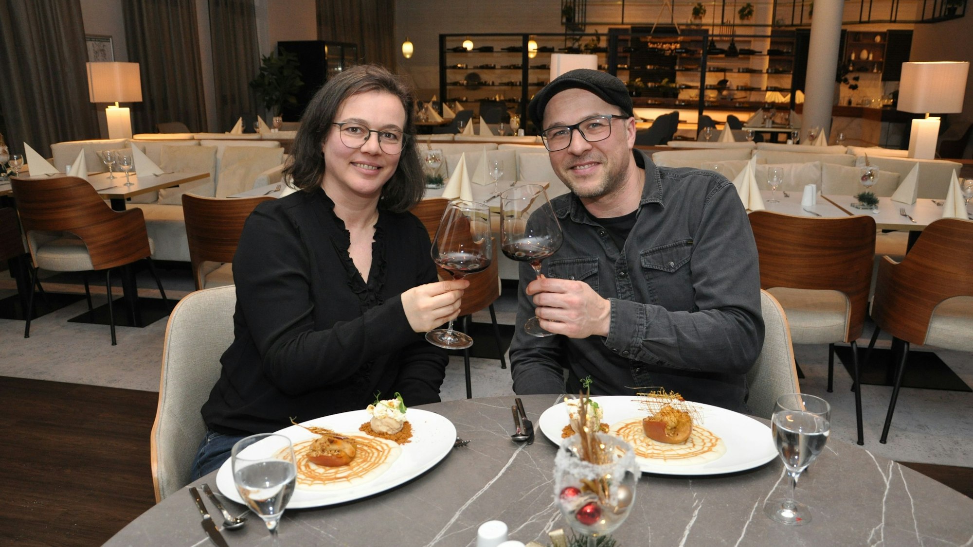 Das Bild zeigt das Paar beim Anstoßen mit Weingläsern. Vor sich haben die beiden einen Teller mit feinem Essen.