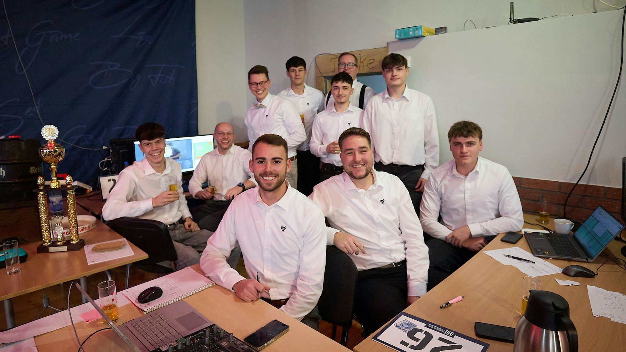 Mehrere Männern in weißen Hemden sitzen und stehen in einem Zimmer und blicken in die Kamera.