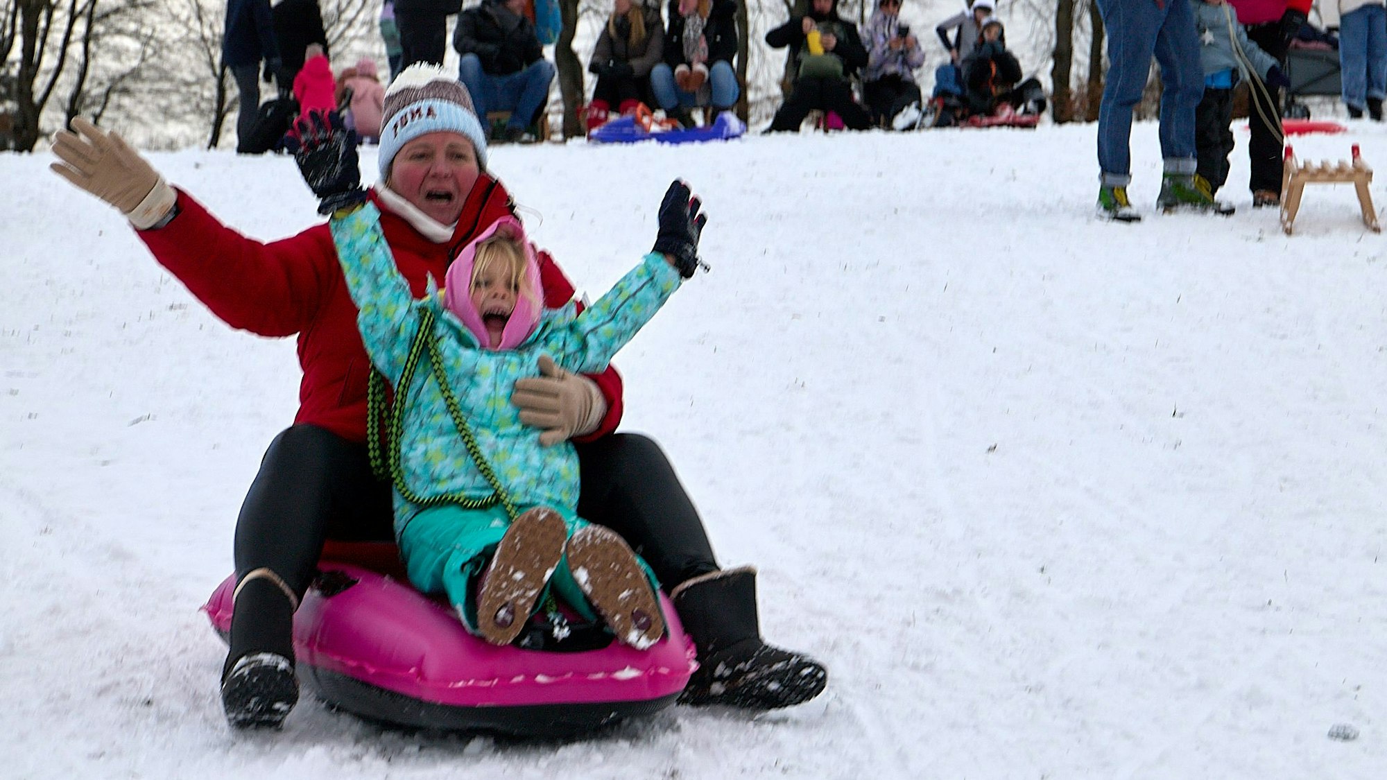 Mutter und Tochter fahren gemeinsam auf einem Schlitten und strecken die Arme in die Höhe.