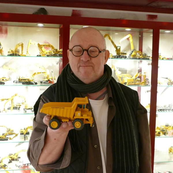 Ein Mann mit Glatze und runder Brille trägt ein braunes Hemd und einen Schal. In seiner rechten Hand hält er ein Modell eines gelben Muldenkippers der Firma Caterpillar.