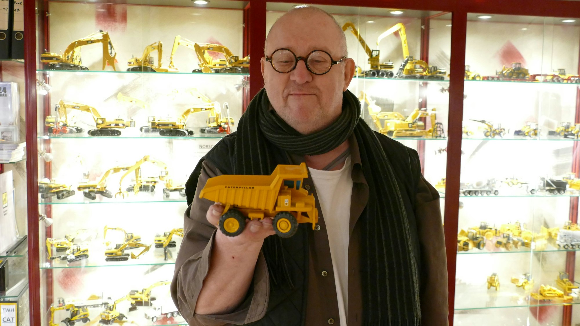 Ein Mann mit Glatze und runder Brille trägt ein braunes Hemd und einen Schal. In seiner rechten Hand hält er ein Modell eines gelben Muldenkippers der Firma Caterpillar.