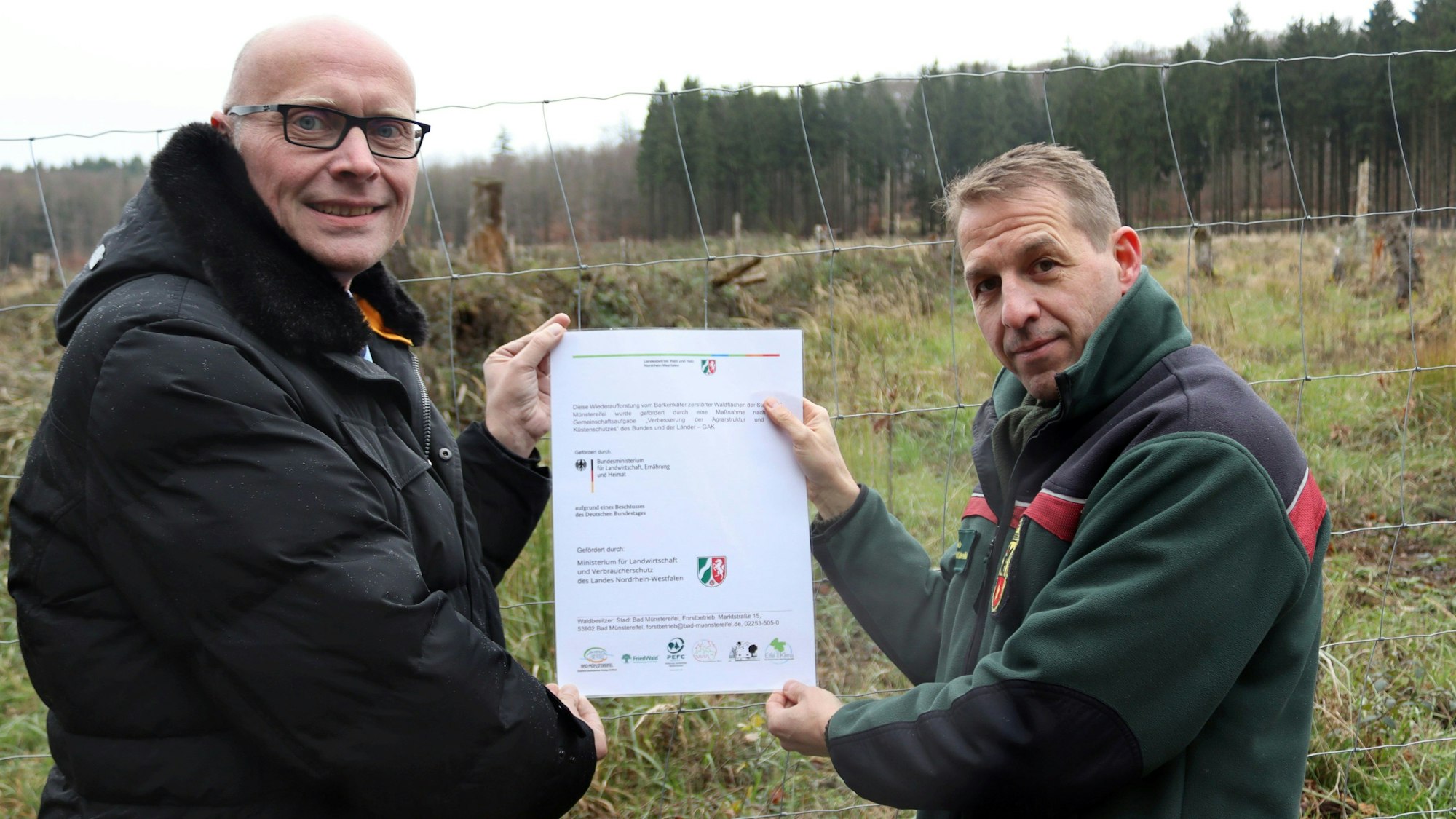 Bürgermeister Sebastian Glatzel (l.) und Stefan Lott, Büroleiter des städtischen Forstbetriebs, zeigen vor einer Waldfläche eine Urkunde des Landes NRW.