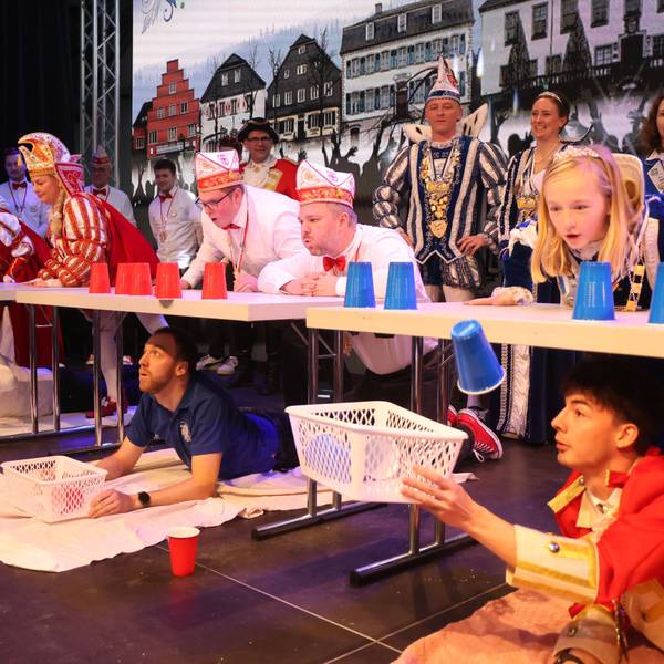 Das Bild zeigt ein kurzweiliges Spiel der Narren: Die einen pusten Plastikbecker über die Tischkante, die anderen liegen bäuchlings unterm Tisch und fangen die Becher mit einem Körbchen auf.