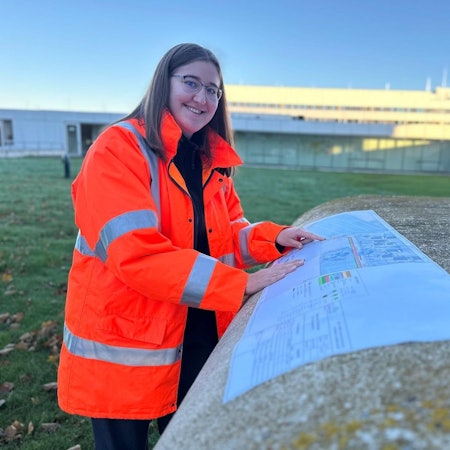 Hannah Bär trägt eine Warnweste und hat ihre Hände auf einen Bauplan gelegt. Sie steht draußen. Im Hintergrund fällt die Sonne auf einen unscharf gestellten Gebäudekomplex.