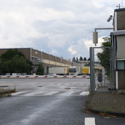 Hinter einem Schlagbaum ist der Betriebshof eines Firmengeländes zu sehen.