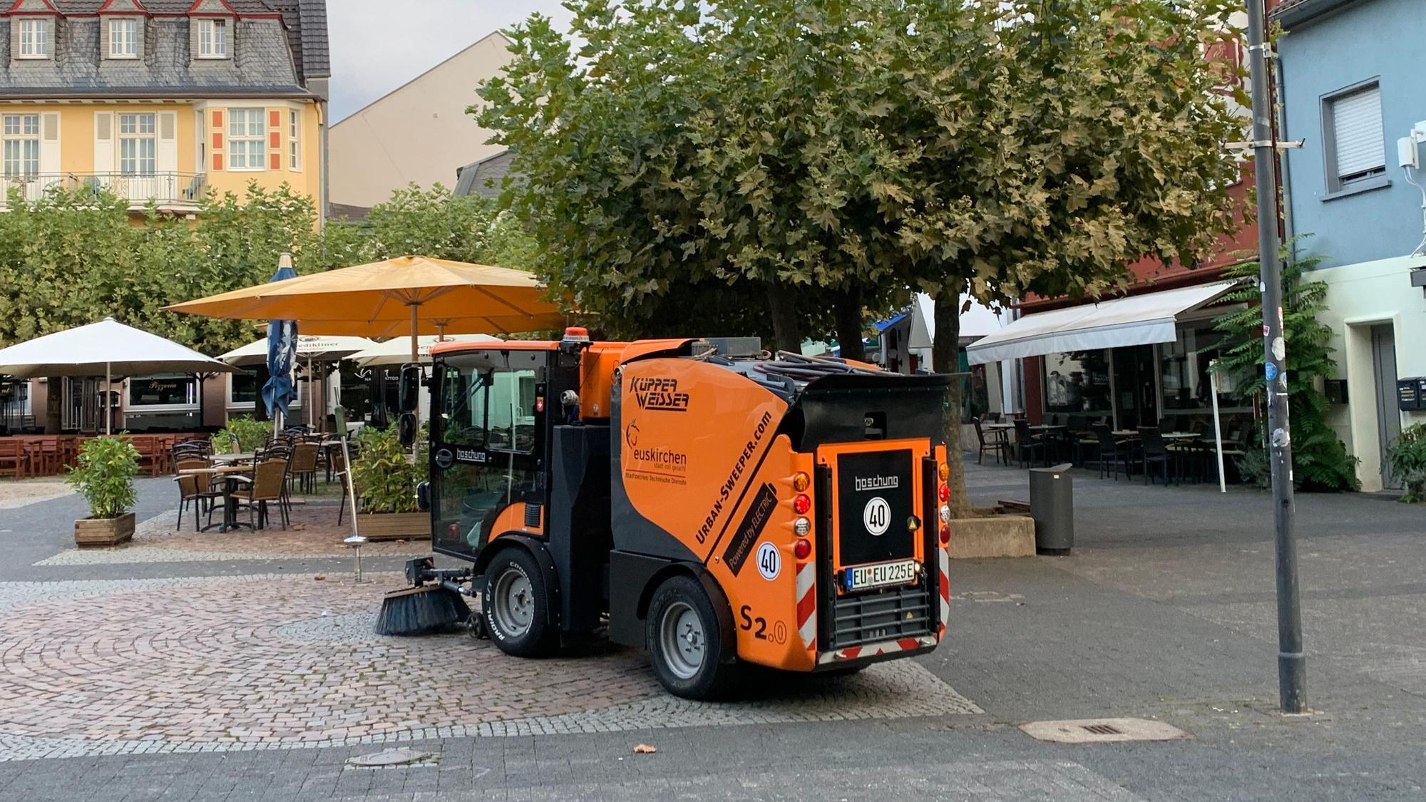 Eine Kehrmaschine reinigt den Platz, im Hintergrund stehen Sonnenschirme.
