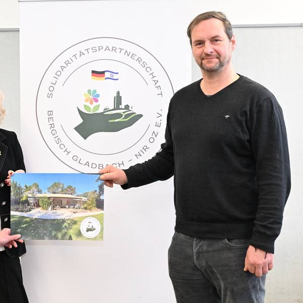 Zwei Männer und eine Frau stehen mit einem Bild von einem Haus in der Hand vor einem Banner, auf dem "Solidaritätspartnerschaftsverein Bergisch Gladbach – Nir Oz" steht.