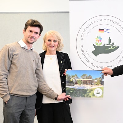 Zwei Männer und eine Frau stehen mit einem Bild von einem Haus in der Hand vor einem Banner, auf dem "Solidaritätspartnerschaftsverein Bergisch Gladbach – Nir Oz" steht.