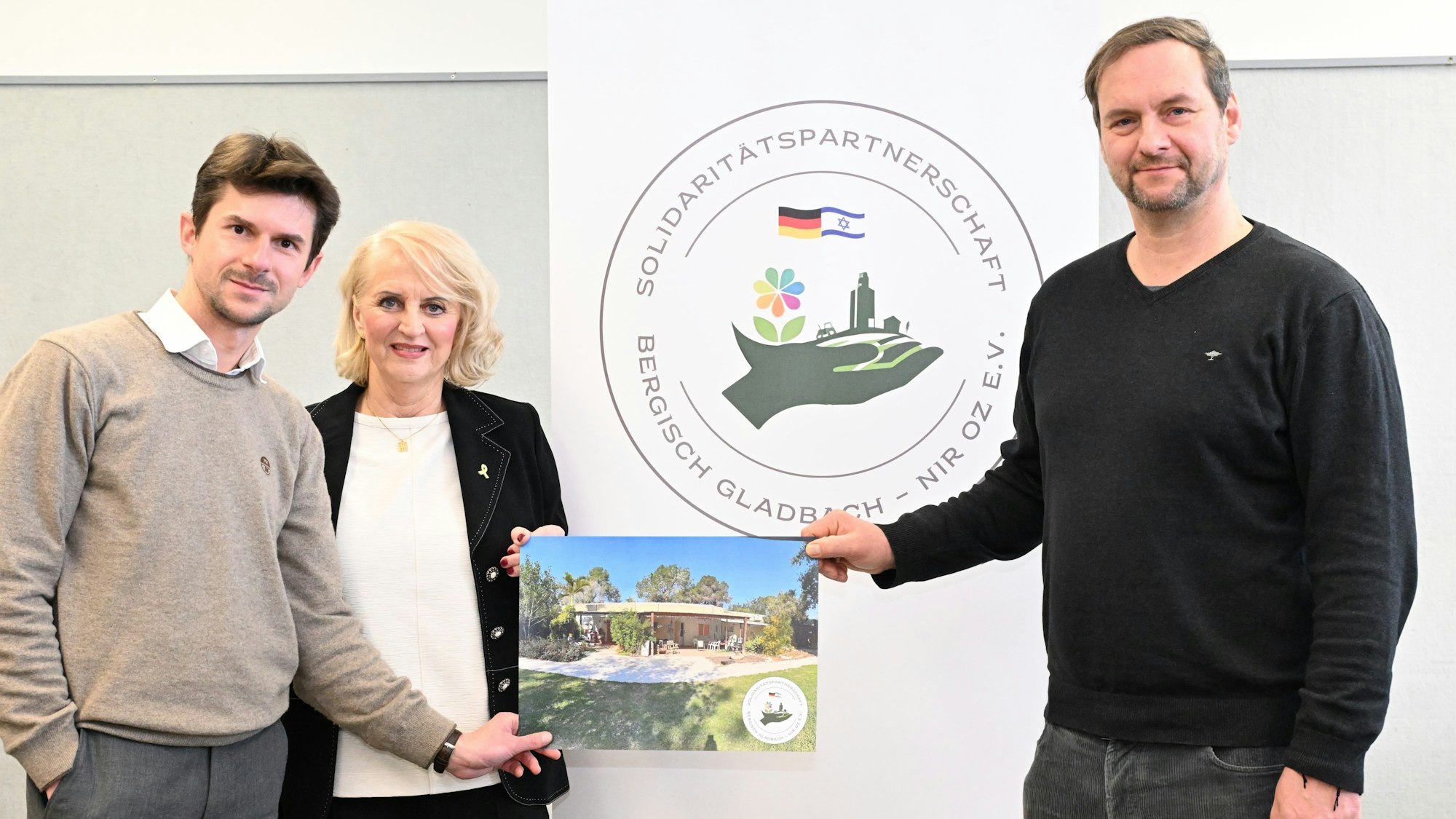 Zwei Männer und eine Frau stehen mit einem Bild von einem Haus in der Hand vor einem Banner, auf dem "Solidaritätspartnerschaftsverein Bergisch Gladbach – Nir Oz" steht.