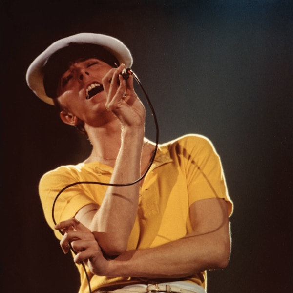 Der etwa 31-jährige David Bowie singt mit geschlossenen Augen und nach hinten geworfenem Kopf in ein Mikrofon. Er trägt ein gelbes Shirt und eine Kapitänsmütze.