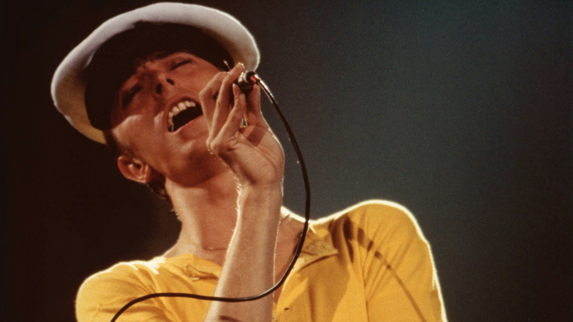 Der etwa 31-jährige David Bowie singt mit geschlossenen Augen und nach hinten geworfenem Kopf in ein Mikrofon. Er trägt ein gelbes Shirt und eine Kapitänsmütze.