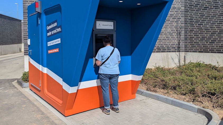 Zu sehen sind der blau-orange Pavillon und ein Kunde am Automaten.