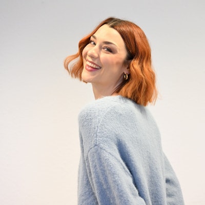 Eine junge Frau mit rotem Haar lächelt über ihre Schulter hinweg in die Kamera.