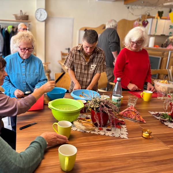 Vier ältere Frauen und ein Mann bereiten an einem Tisch eine Suppe zu. Auf dem Tisch liegen Schalen für geschnittene Zutaten und Schneidebretter.