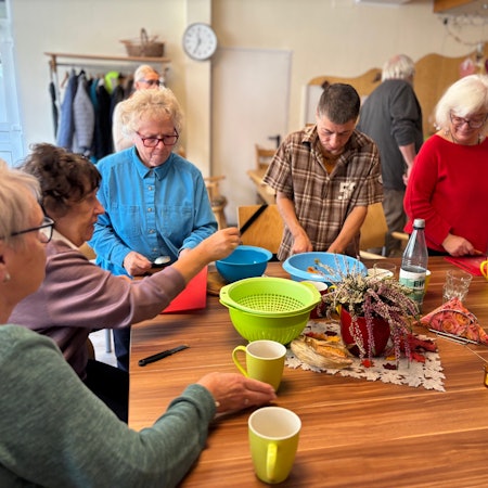 Vier ältere Frauen und ein Mann bereiten an einem Tisch eine Suppe zu. Auf dem Tisch liegen Schalen für geschnittene Zutaten und Schneidebretter.