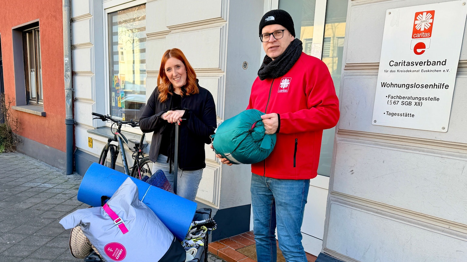 Die beiden Sozialarbeiter der Caritas stehen vor dem Gebäude der Wohnungslosenhilfe. Sie haben einen blauen Bollerwagen dabei, auf dem sich Decken, Schlafsäcke und Isomatten befinden.