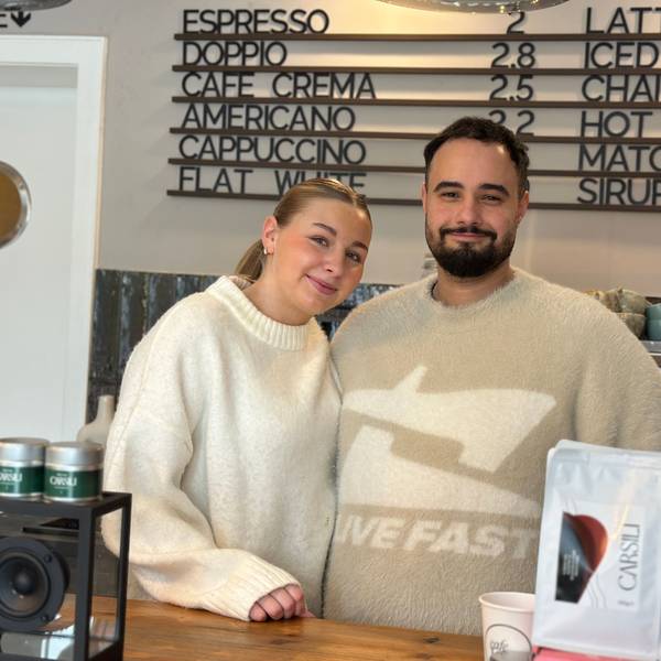 Eine blonde Frau und ein Mann mit dunklem Haar und Bart stehen hinter dem Verkaufstresen eines Cafés.