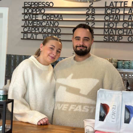 Eine blonde Frau und ein Mann mit dunklem Haar und Bart stehen hinter dem Verkaufstresen eines Cafés.