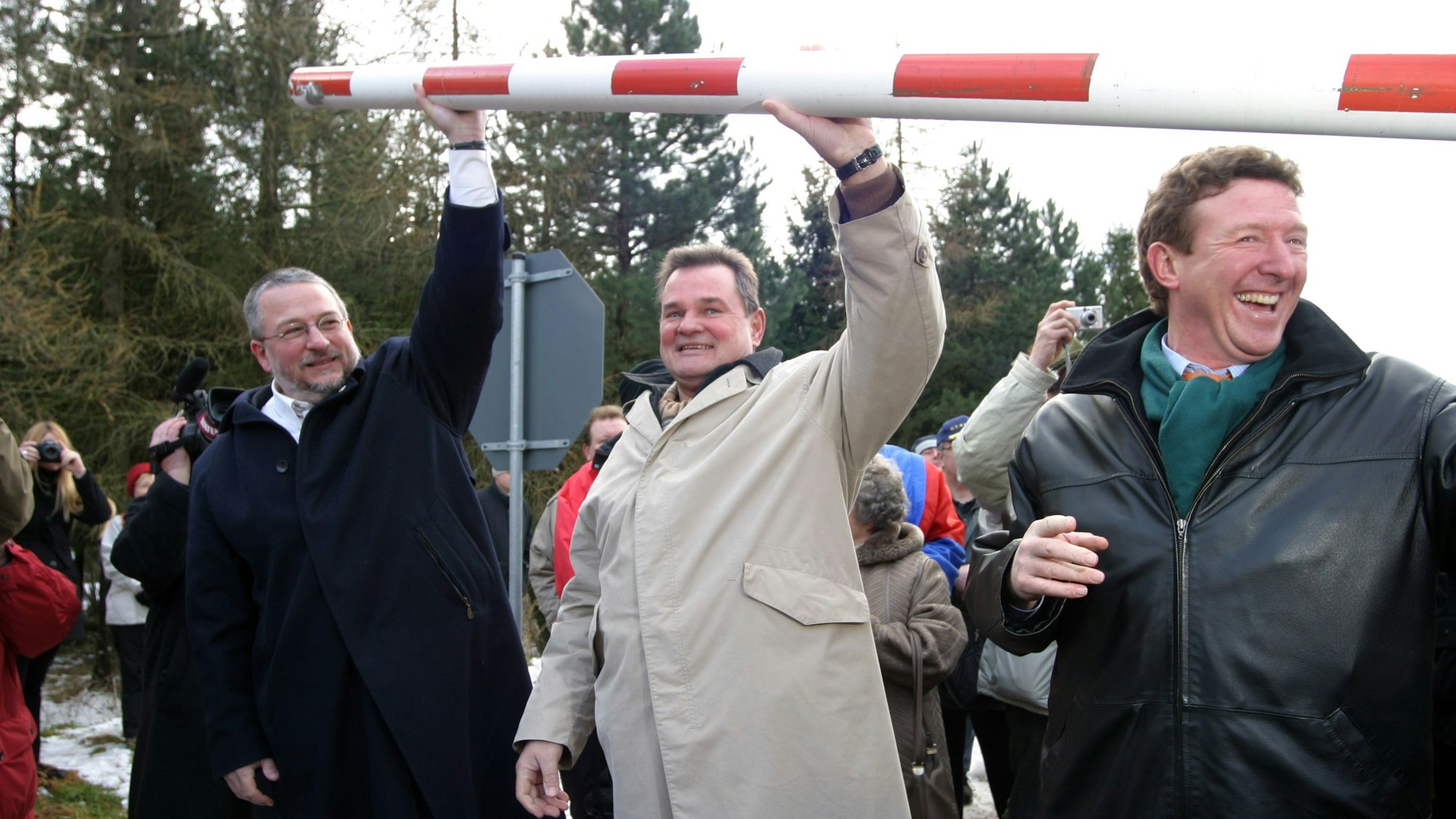Ralf Hergarten, Günter Rosenke und Manfred Poth heben den Schlagbaum über ihre Köpfe.Im Hintergrund sind andere Menschen zu sehen.