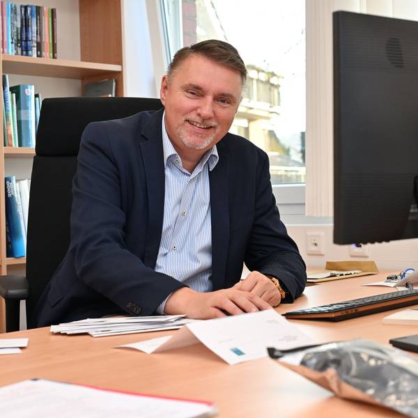 Ein Mann mittleren Alters sitzt an einem Schreibtisch und lächelt.