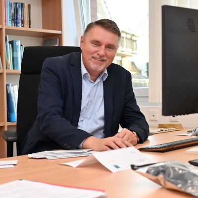 Ein Mann mittleren Alters sitzt an einem Schreibtisch und lächelt.