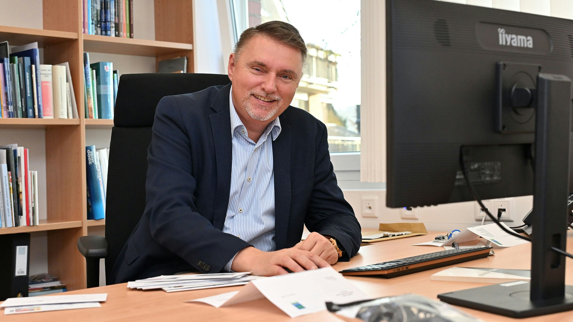 Ein Mann mittleren Alters sitzt an einem Schreibtisch und lächelt.