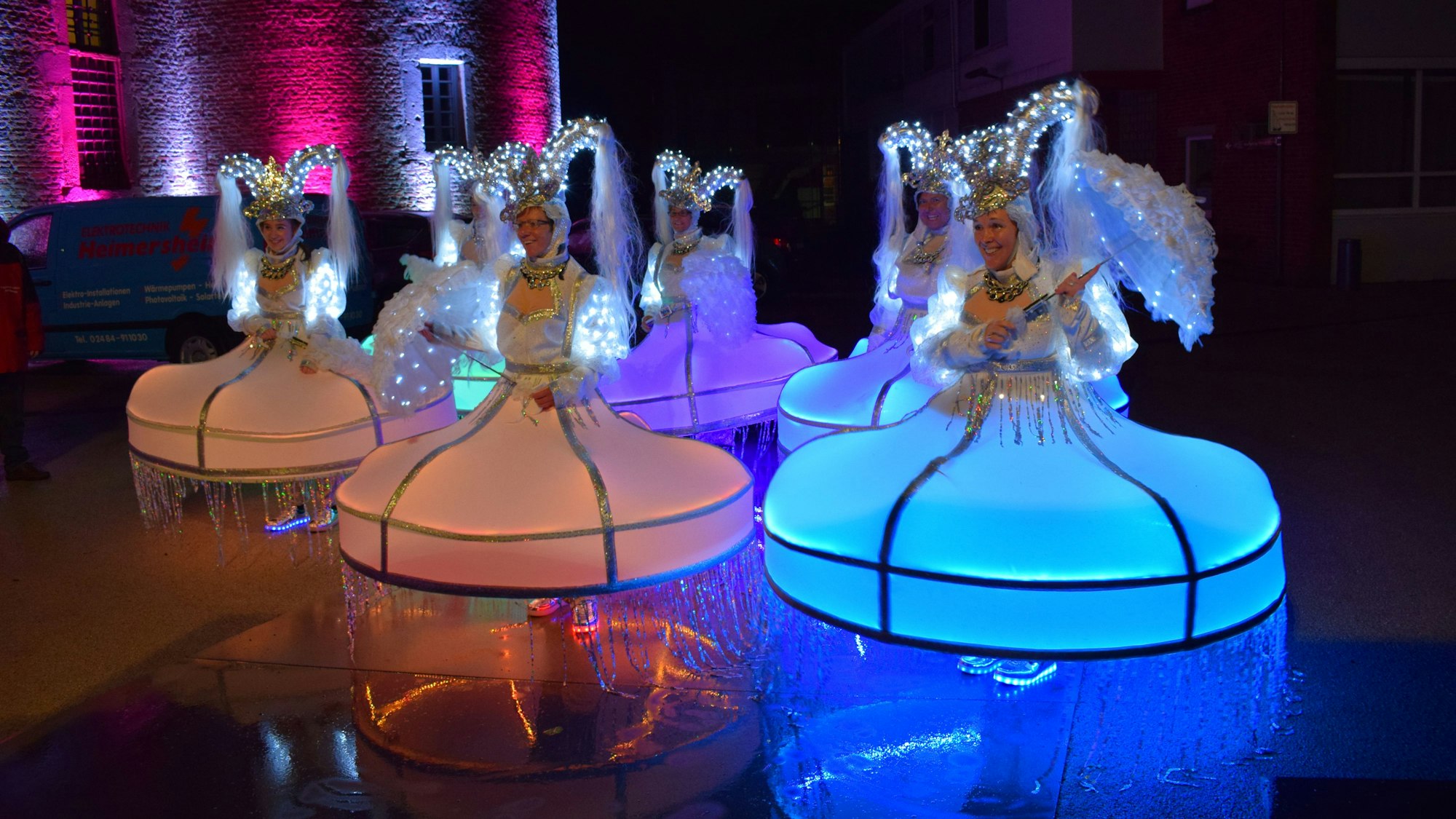 Frauen in weit ausfallenden Kostümen, die an pompöse Kleider erinnern, stehen auf einer glatten Fläche, in der sich die leuchtenden Anziehsachen spiegeln.