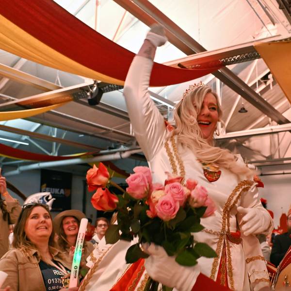 Prinzessin Colette I. in einem weißen Prinzenkleid beim Einzug in den Saal. Rechts steht ein Adjutant, der ihr einen Strauß Rosen anreicht.