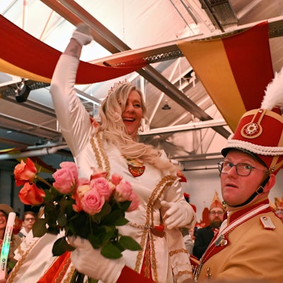 Prinzessin Colette I. in einem weißen Prinzenkleid beim Einzug in den Saal. Rechts steht ein Adjutant, der ihr einen Strauß Rosen anreicht.