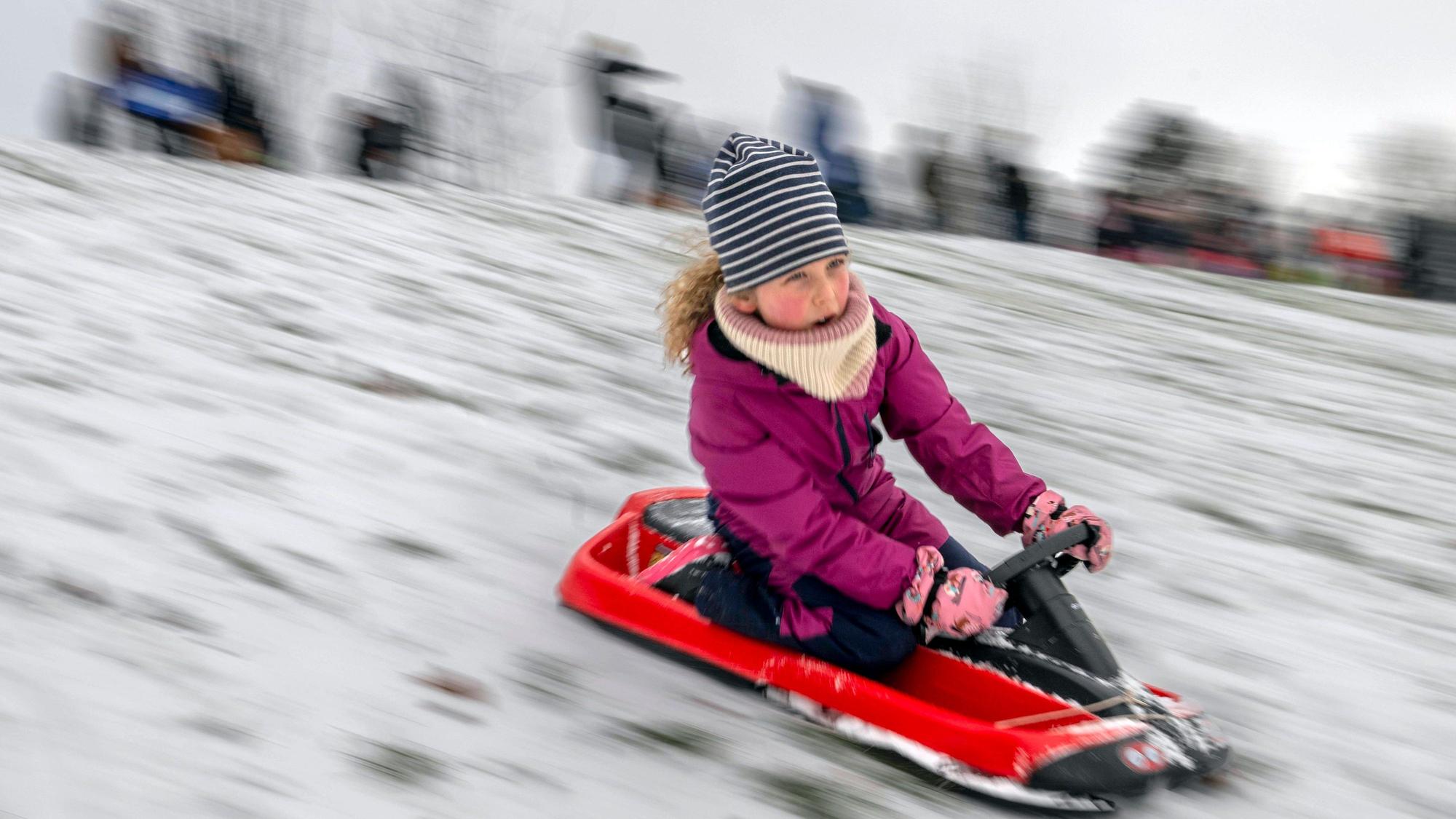 Ein Mädchen mit Mütze, Schal und lilafarbener Winterjacke rodelt auf einem roten Schlitten einen Hang hinunter.