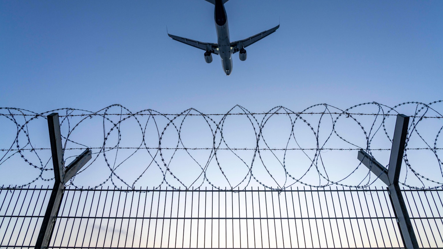 Symbolbild Sicherheit auf dem Flughafen, äußerer Zaun am Airport Düsseldorf International, Stahldrahtzaun, mit S-Draht Rollen, NATO-Draht, auf der Zaunkrone, Flugzeug im Landeanflug, Sonnenuntergang, NRW, Deutschland, Sicherheit Flughafen *** Symbolic image security at the airport, outer fence at Düsseldorf International Airport, steel wire fence, with S wire rolls, NATO wire, on the fence top, airplane approaching, sunset, NRW, Germany, airport security