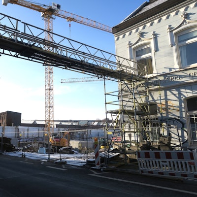 An einer Straße ist eine große Baulücke. Ein Kran und Baugerüste stehen dort hinter einem Bauzaun.
