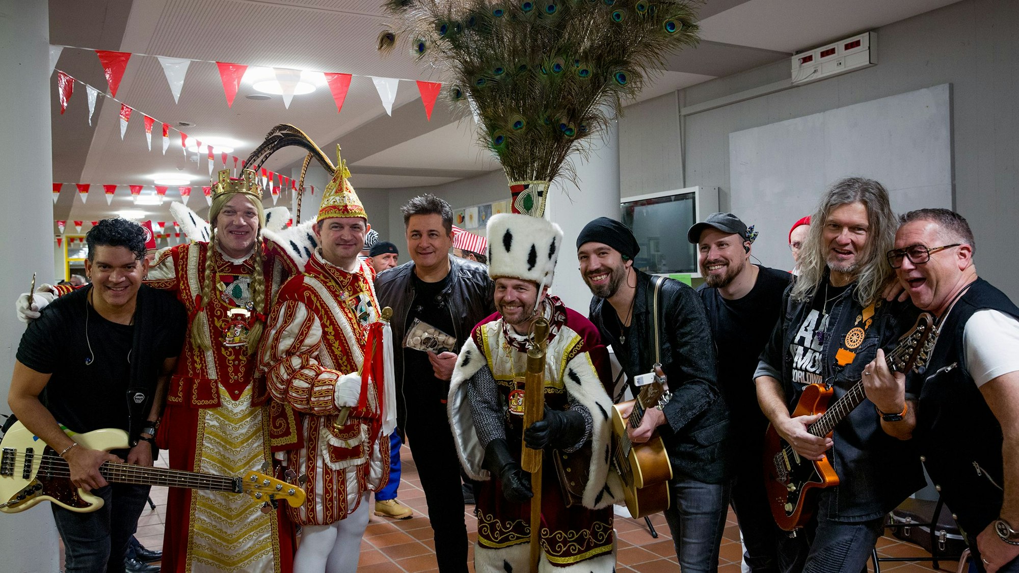 Mehrere Menschen stehen in einem Schulflur, darunter ein als Dreigestirn kostümiertes Ensemble und Mitglieder einer Band, teils mit Gitarren.