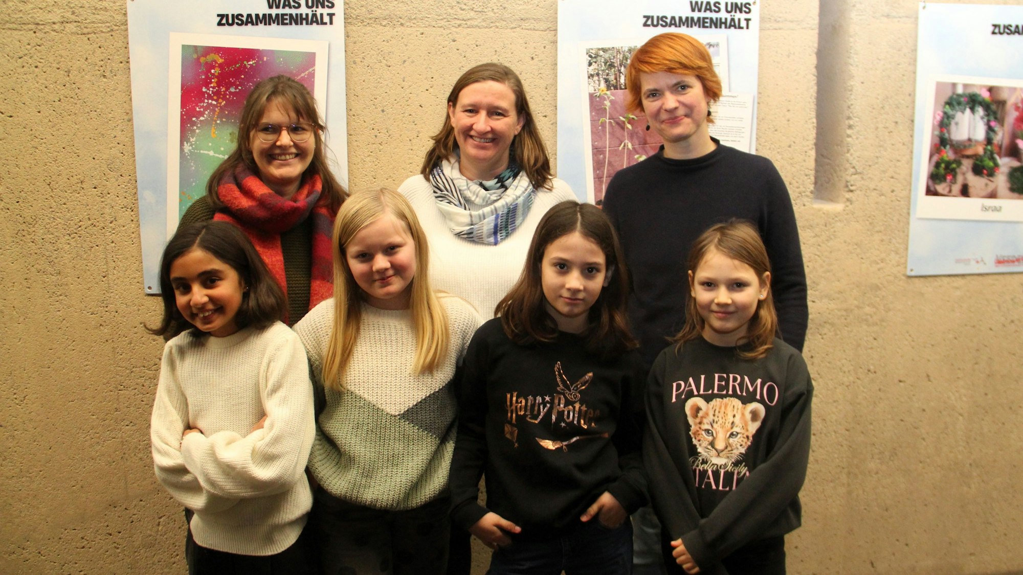 Drei Frauen und vier Mädchen stehen zusammen vor einer Wand, auf der Plakate mit der Aufschrift "Was uns zusammenhält" zu sehen sind.