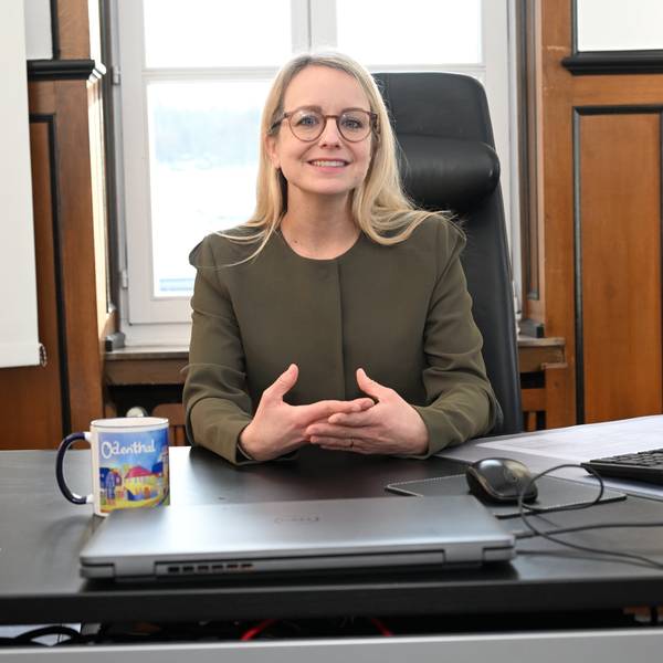Eine Frau mit blondem, langen Haar und Brille sitzt an einem Schreibtisch.