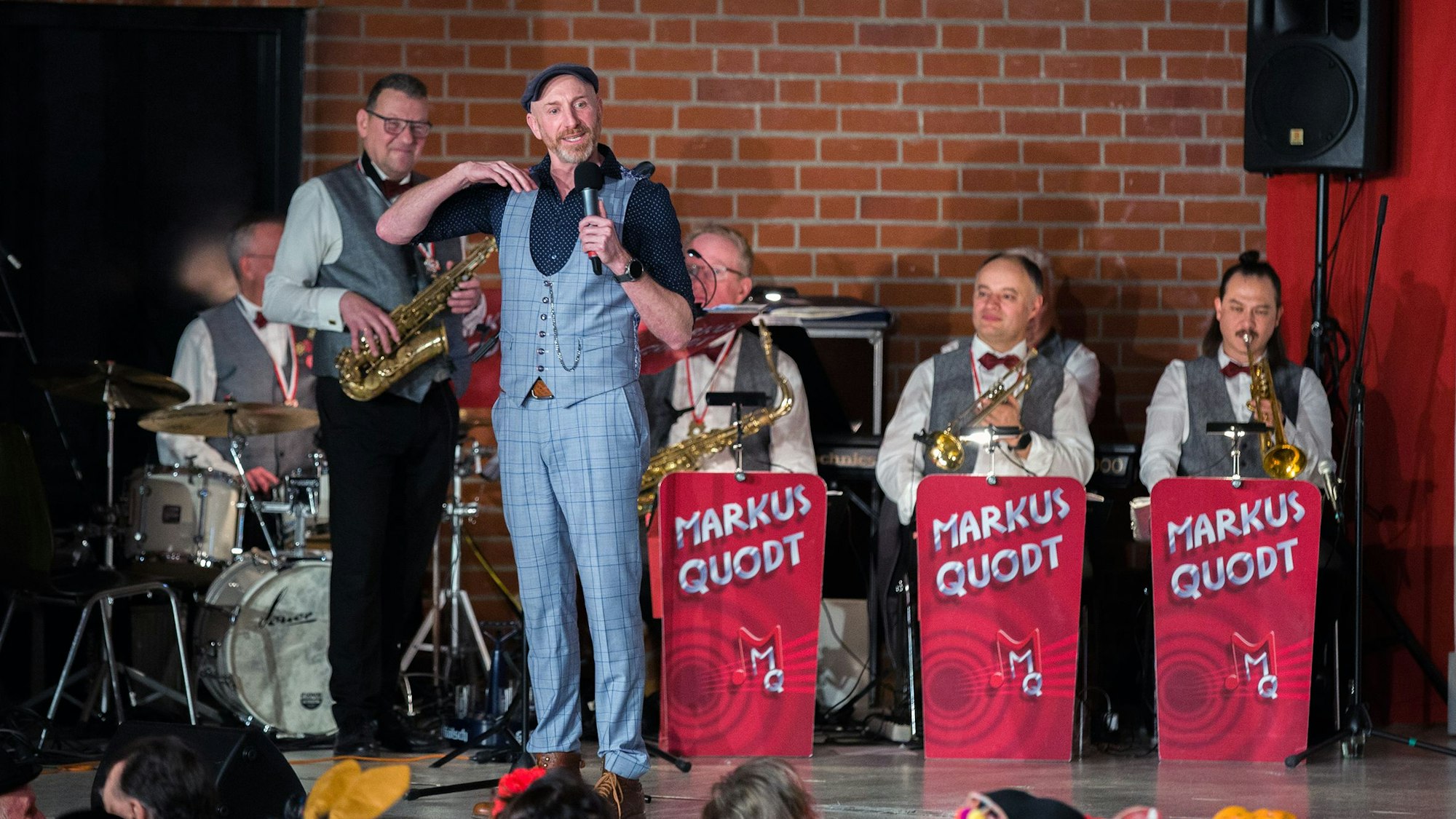Ein Mann mit Kappe steht auf einer Bühne, hinter ihm sitzen Musiker mit Trompeten und Saxophon.