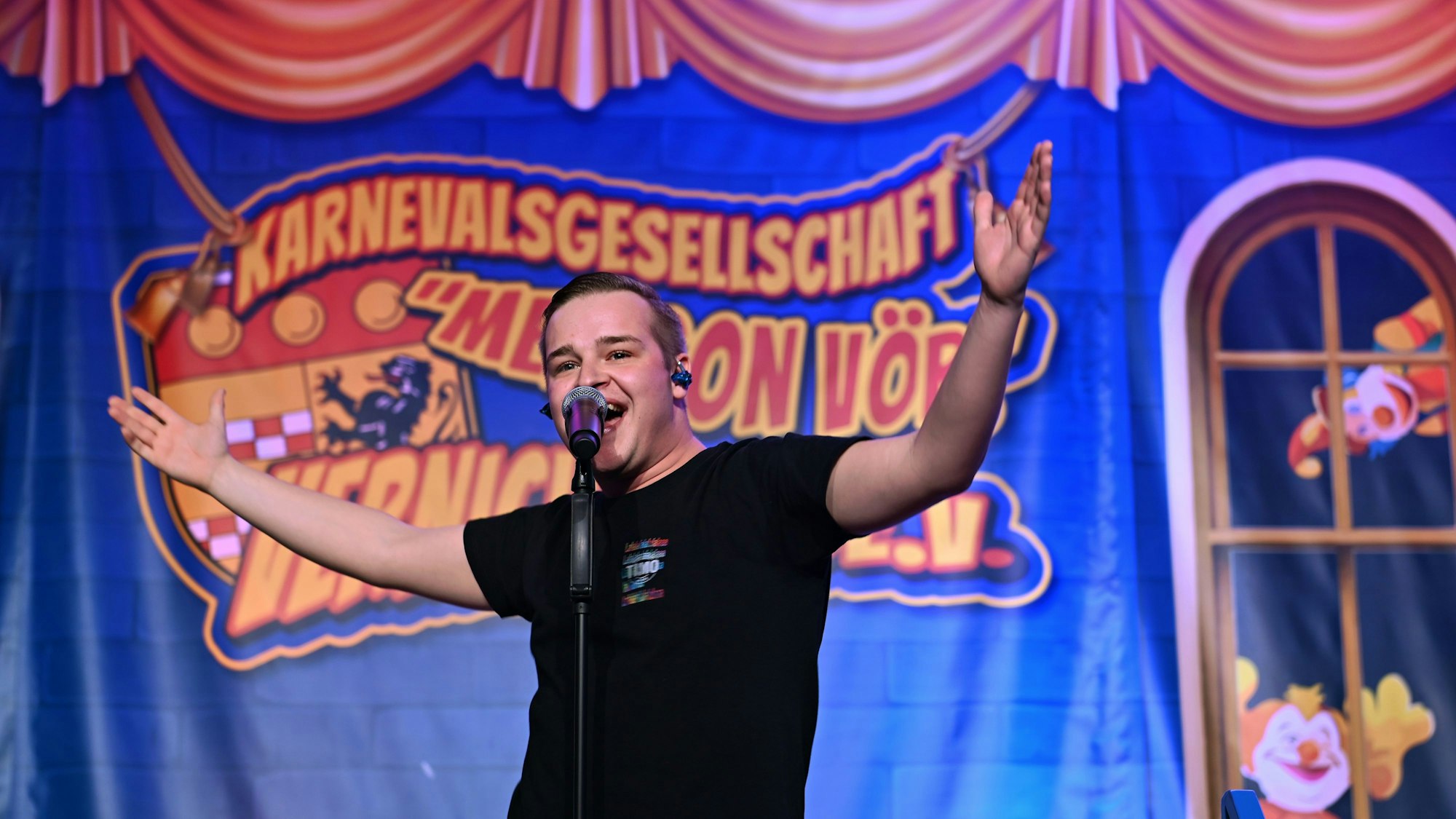 Timo Schwarzendahl hat die Arme weit von sich gestreckt und spricht in ein Mikrofon auf einem Mikrofonständer.