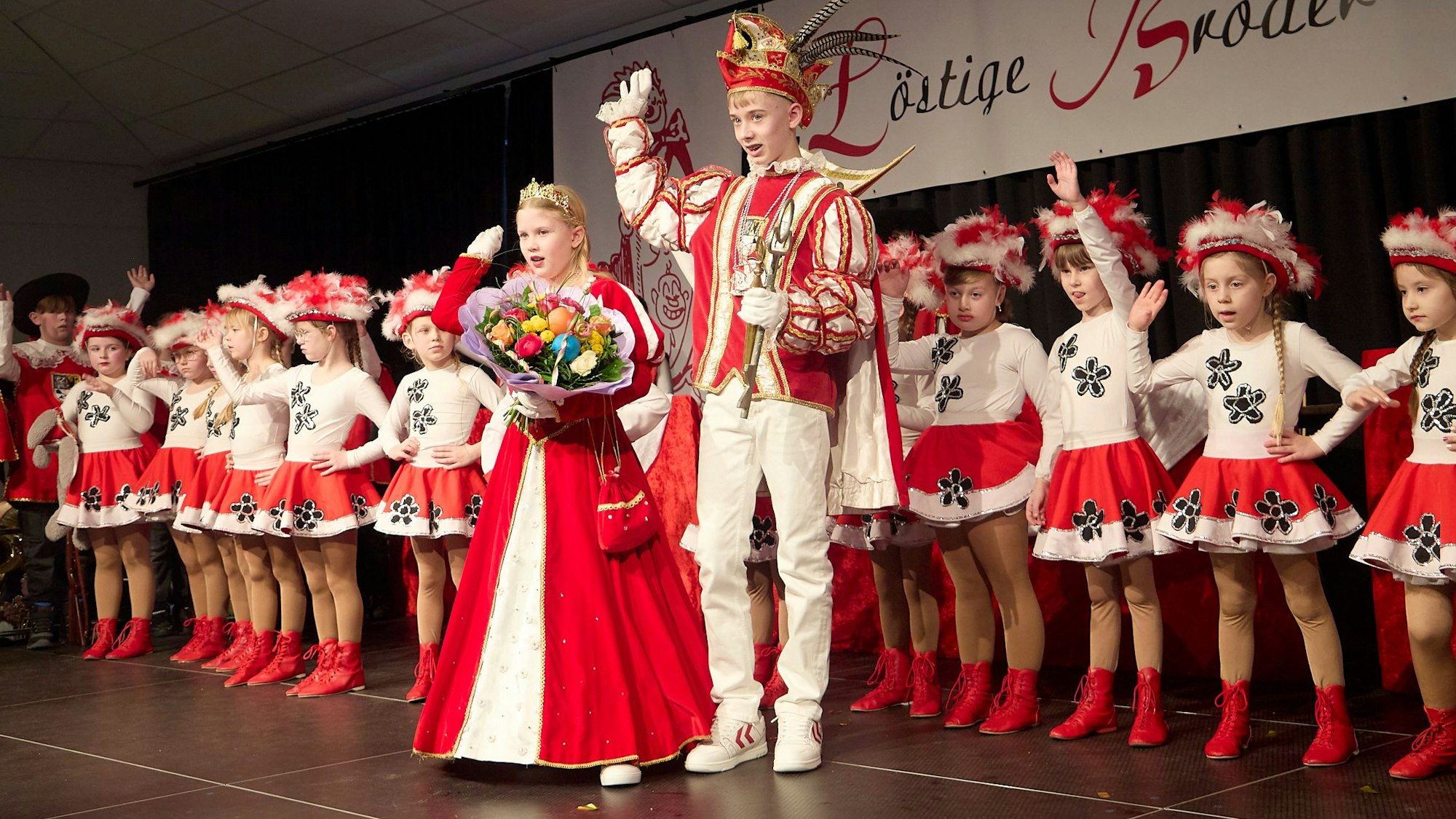 Auf der Bühne stehen Kinder der Tanzgarden der „Löstige Bröder“. Davor steht das Prinzenpaar und hebt die Arme zum Alaaf-Ruf in die Höhe.