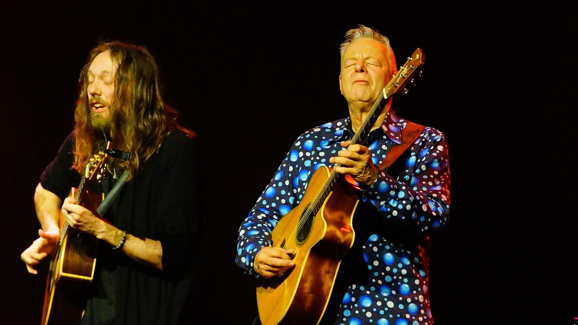 Das Bild zeigt Mike Dawes und Tommy Emmanuel. Beide spielen Gitarre. Der Hintergrund ist schwarz.