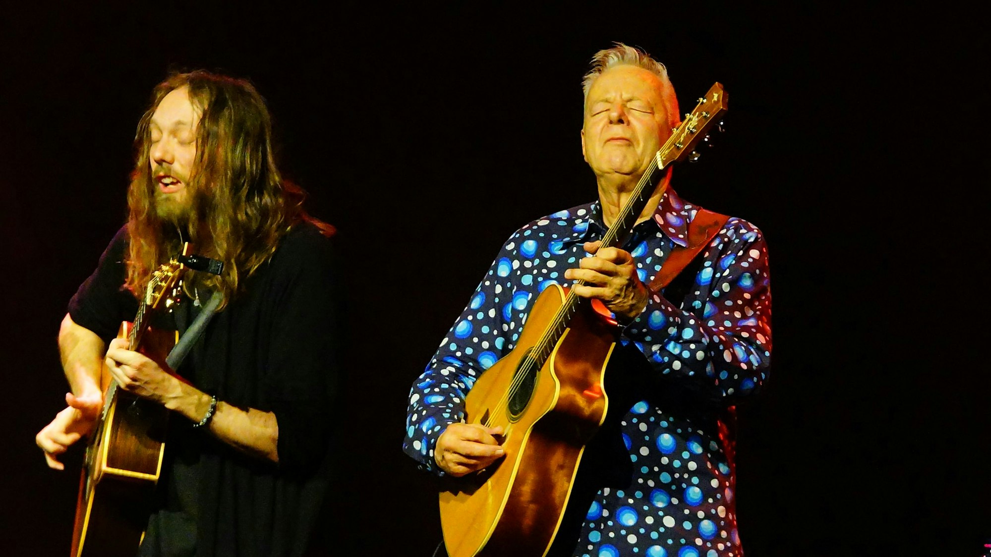 Das Bild zeigt Mike Dawes und Tommy Emmanuel. Beide spielen Gitarre. Der Hintergrund ist schwarz.