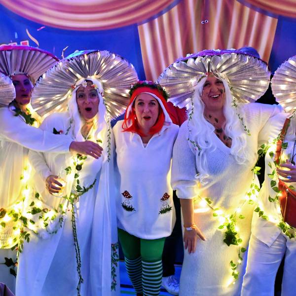 Sechs Frauen in Waldpilzkostümen, die teilweise beleuchtet sind, posieren fröhlich für die Kamera.