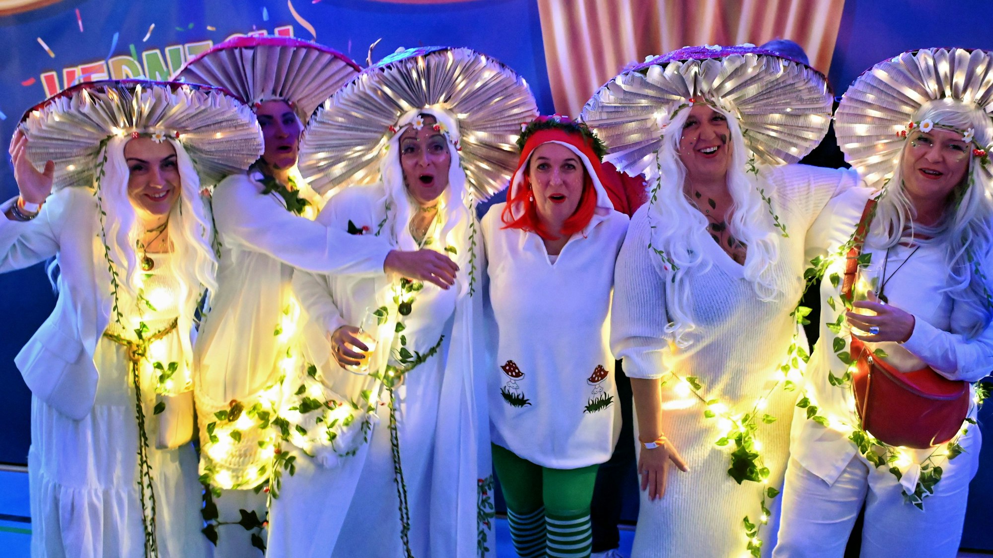 Sechs Frauen in Waldpilzkostümen, die teilweise beleuchtet sind, posieren fröhlich für die Kamera.