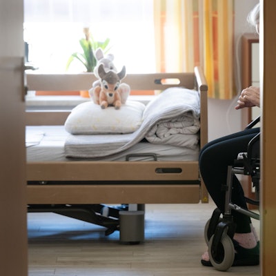 Ein Bett im Heim, darauf Kuscheltiere, daneben sitzt eine Person im Rollstuhl.