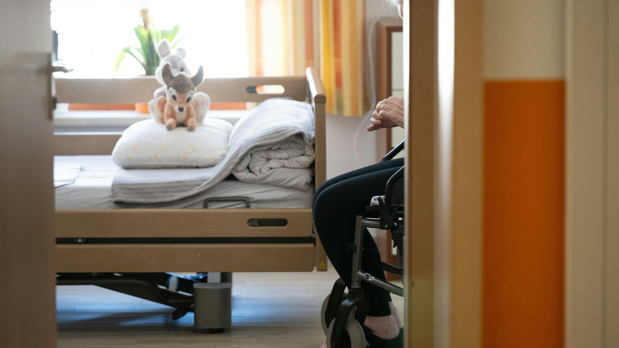 Ein Bett im Heim, darauf Kuscheltiere, daneben sitzt eine Person im Rollstuhl.