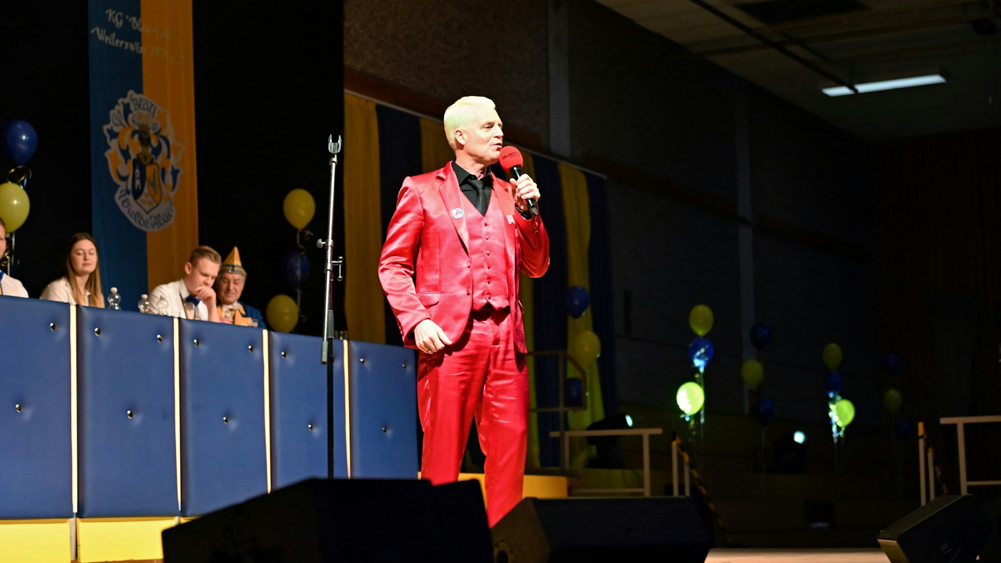 Guido Cantz trägt einen knalligen, roten Anzug und spricht auf eine Bühne in ein Mikrofon.