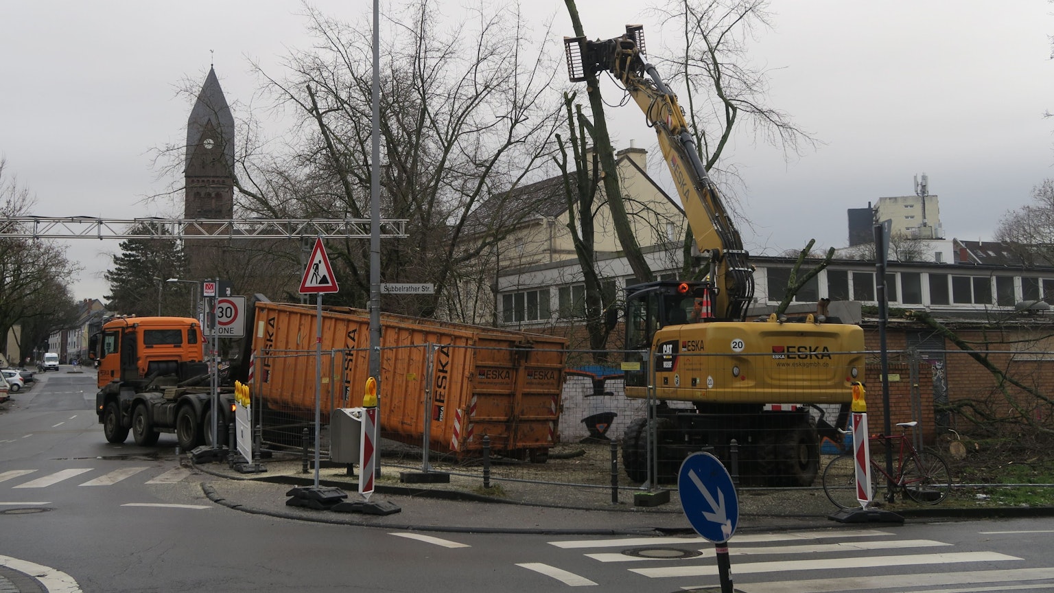 An einer Straßenecke ist ein Bauzaun, ein Lkw und ein Bagger sind dahinter zugange.