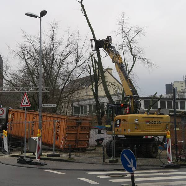 An einer Straßenecke ist ein Bauzaun, ein Lkw und ein Bagger sind dahinter zugange.