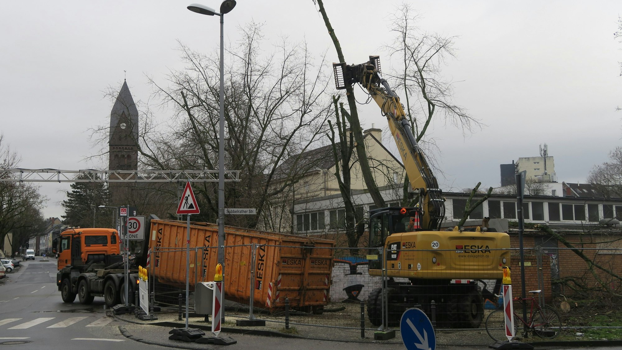 An einer Straßenecke ist ein Bauzaun, ein Lkw und ein Bagger sind dahinter zugange.