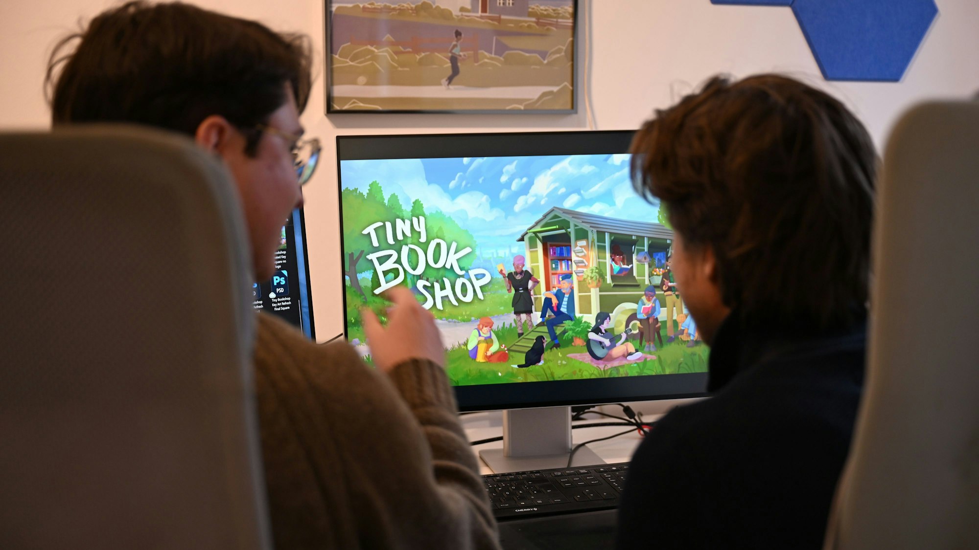 Die Computerspiele-Entwickler David Zapfe-Wildemann und Raven Rusch von dem Kölner Independent-Gamingstudio Neoludic sitzen in ihrem Büro vor einem Bildschirm, auf dem die Startseite des Spiels Tiny Bookshop zu sehen ist.